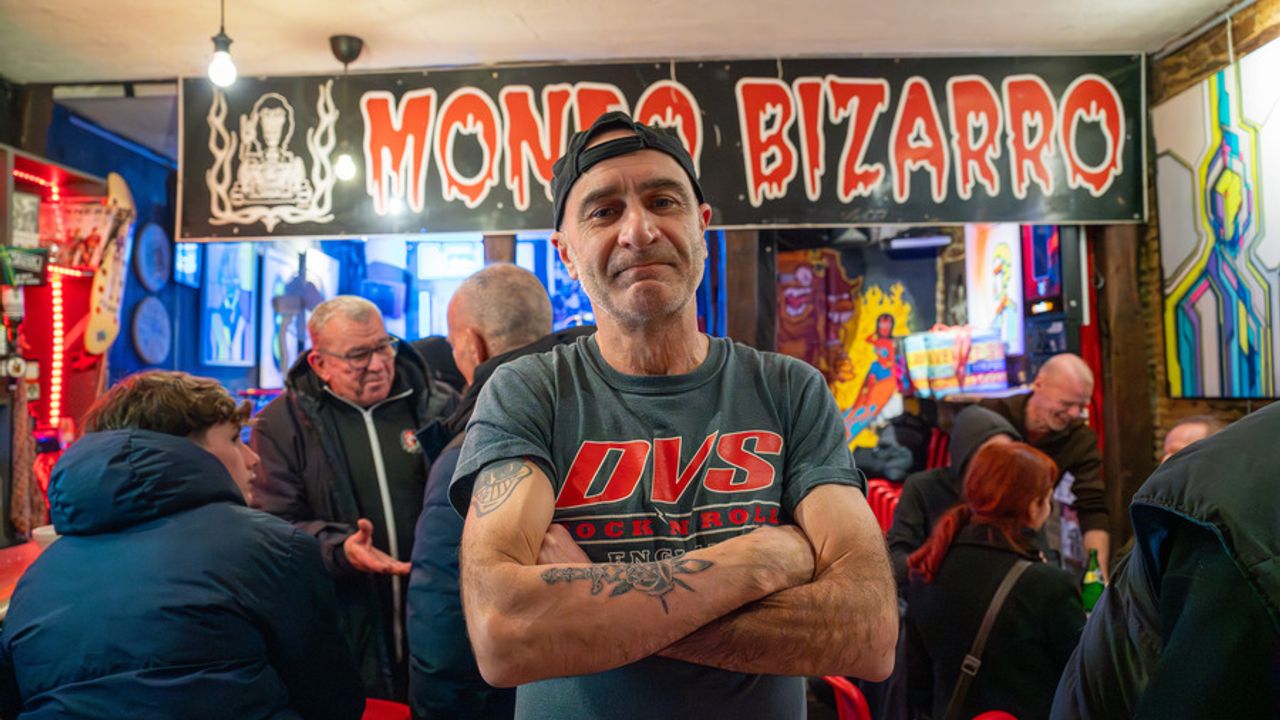 Bruno Perrin bras croisés dans le bar la Trinquette. Derrière lui des clients et au dessus de lui l'ensiegne du Mondo Bizarro.