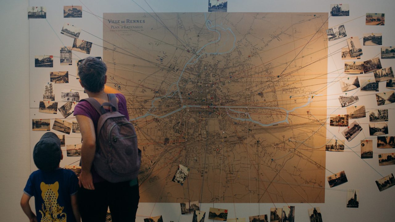 Une grande carte de la ville accrochée au mur est reliée par des fils à des cartes postales anciennes. Une femme et un enfant regardent ce travail de cartographie.