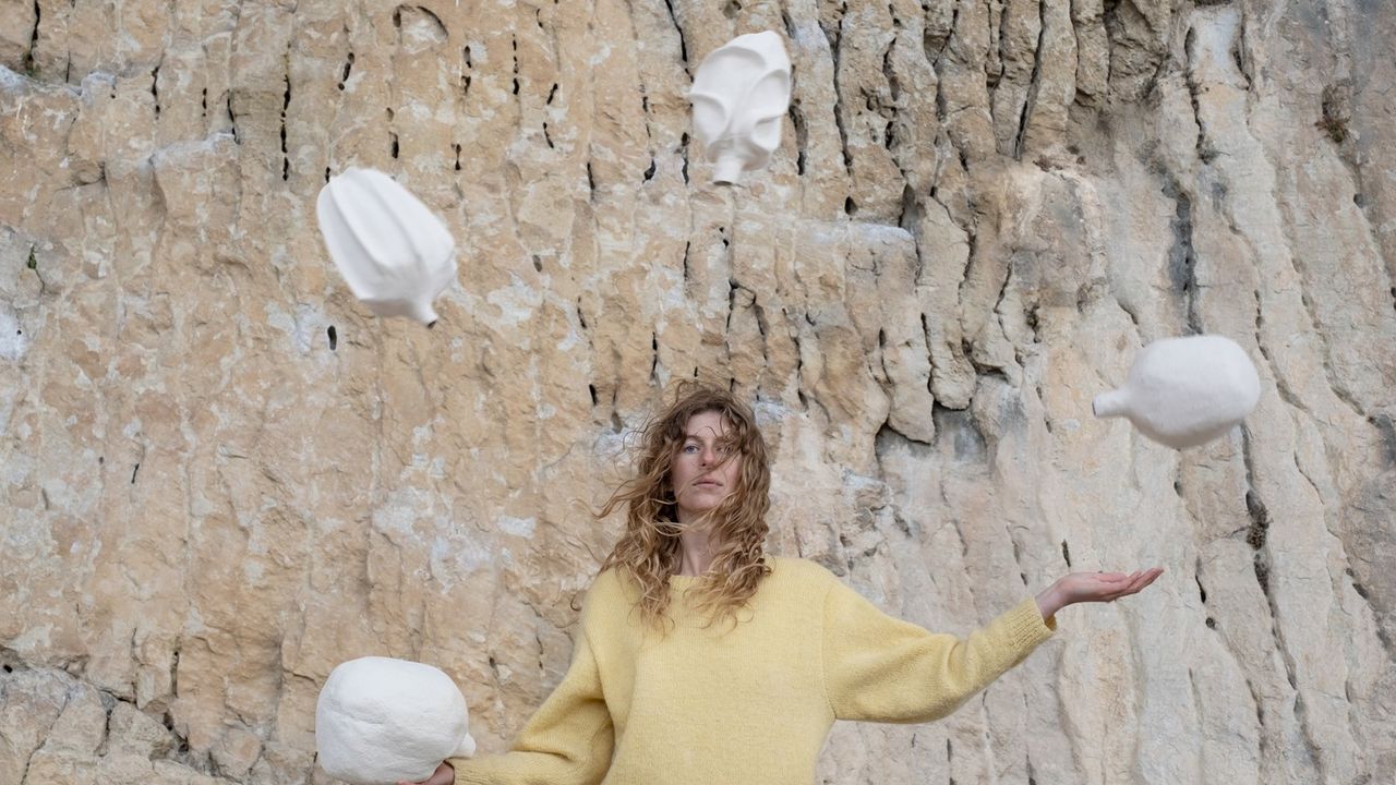 On voit l'artiste jongler avec des vases en céramique devant une falaise rocheuse. Le vent agite ses cheveux qui recouvrent partiellement son visage.