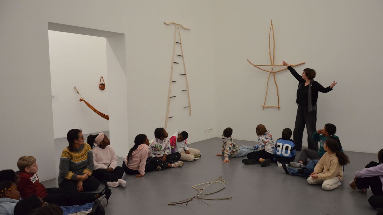 Des enfants sont assis en tailleur au sein de l'exposition dansetremblenage d'Hélène Bertin. Ils regardent tous la médiatrice culturelle Amandine Braud, qui interprète avec ses bras et son corps les sculptures d'Hélène Bertin.