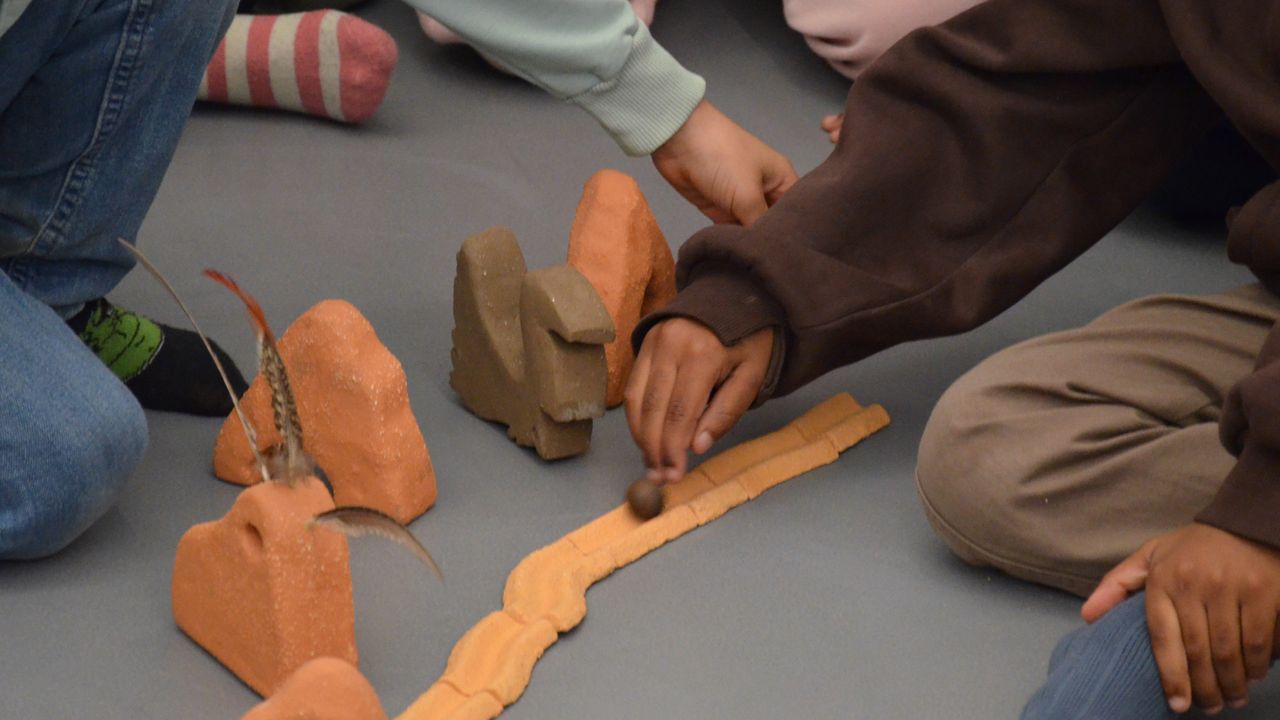 On voit une route formée par plusieurs tessons de céramique au sol. On aperçoit des objets en céramique brute et ocre, ils ressemblent à des montagnes. L'un est orné de trois plumes. Un autre est plus foncé et représente un renne. Des enfants poussent une bille de galle le long de la route.