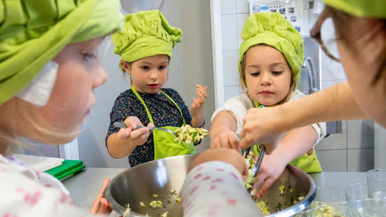 Vêtus de toques et de tabliers, des enfants préparent un plat en cuisine. 
