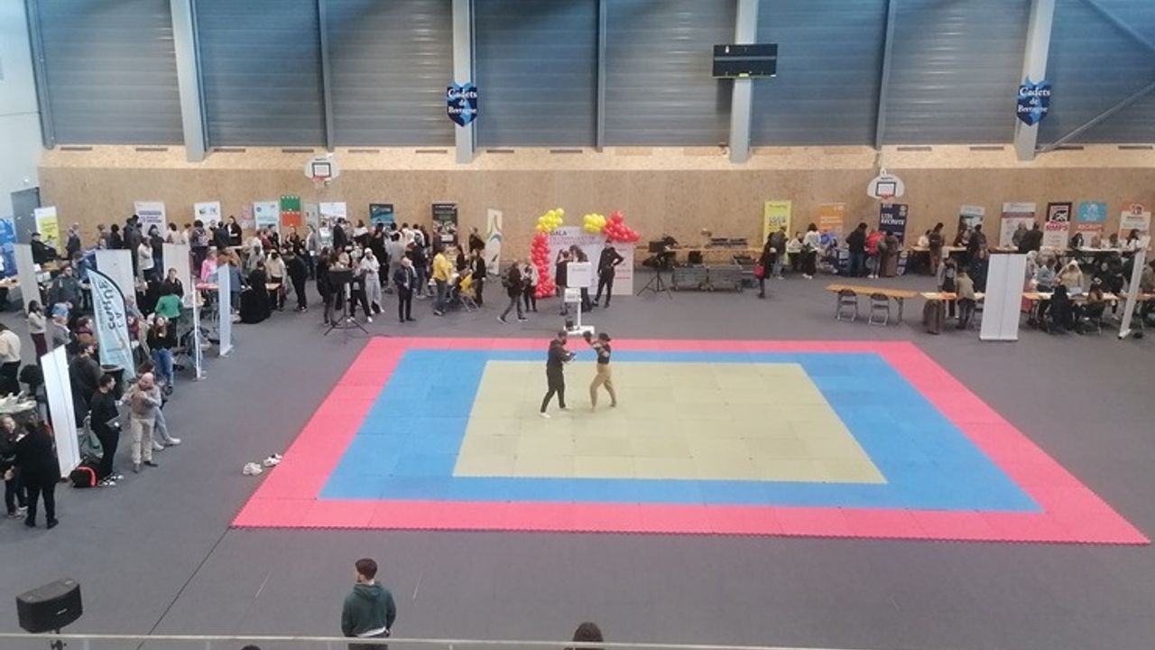 Deux personnes sur un tatami au centre d'un gymnase avec des stands tout autour.