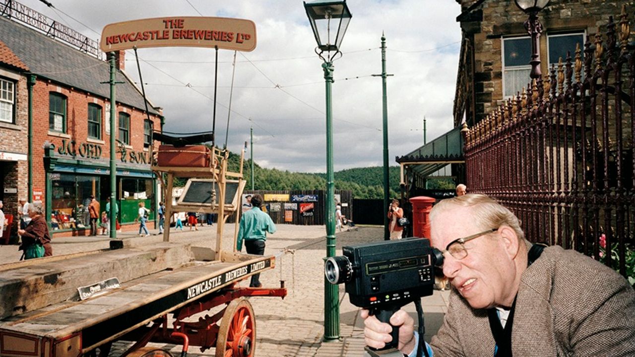 Au premier plan, un homme regarde dans le viseur d'une caméra. Il tourne le dos à la scène composée notamment d'un charriot évoquant un décor de western.