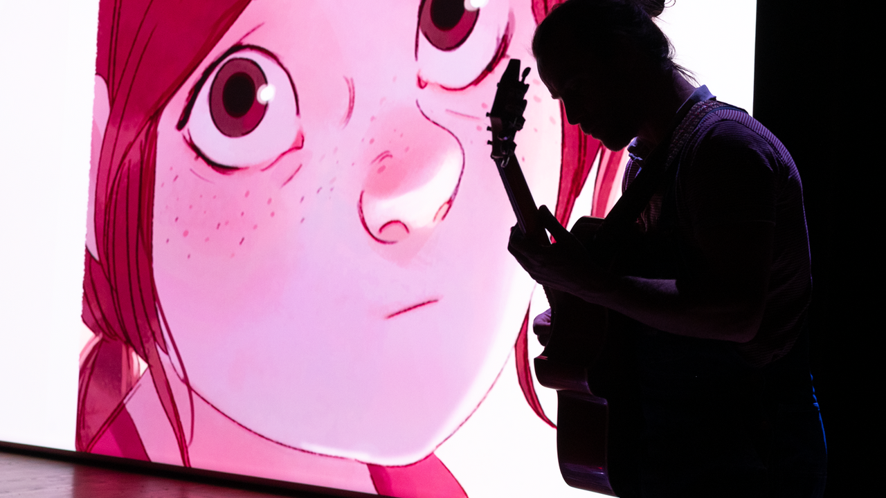 Au premier plan, un homme joue de la guitare avec un visage rose dessiné projeté sur un écran au second plan