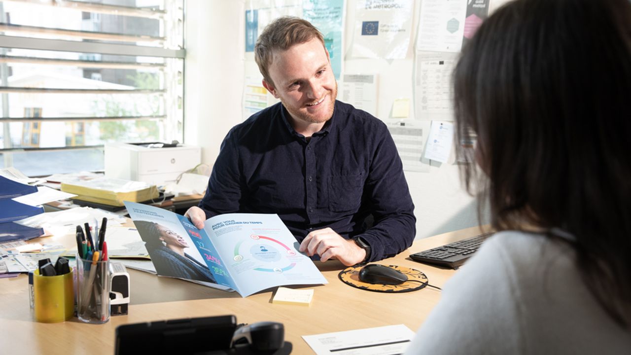 Un homme assis en face d’une femme à un bureau, en train de montrer un document.