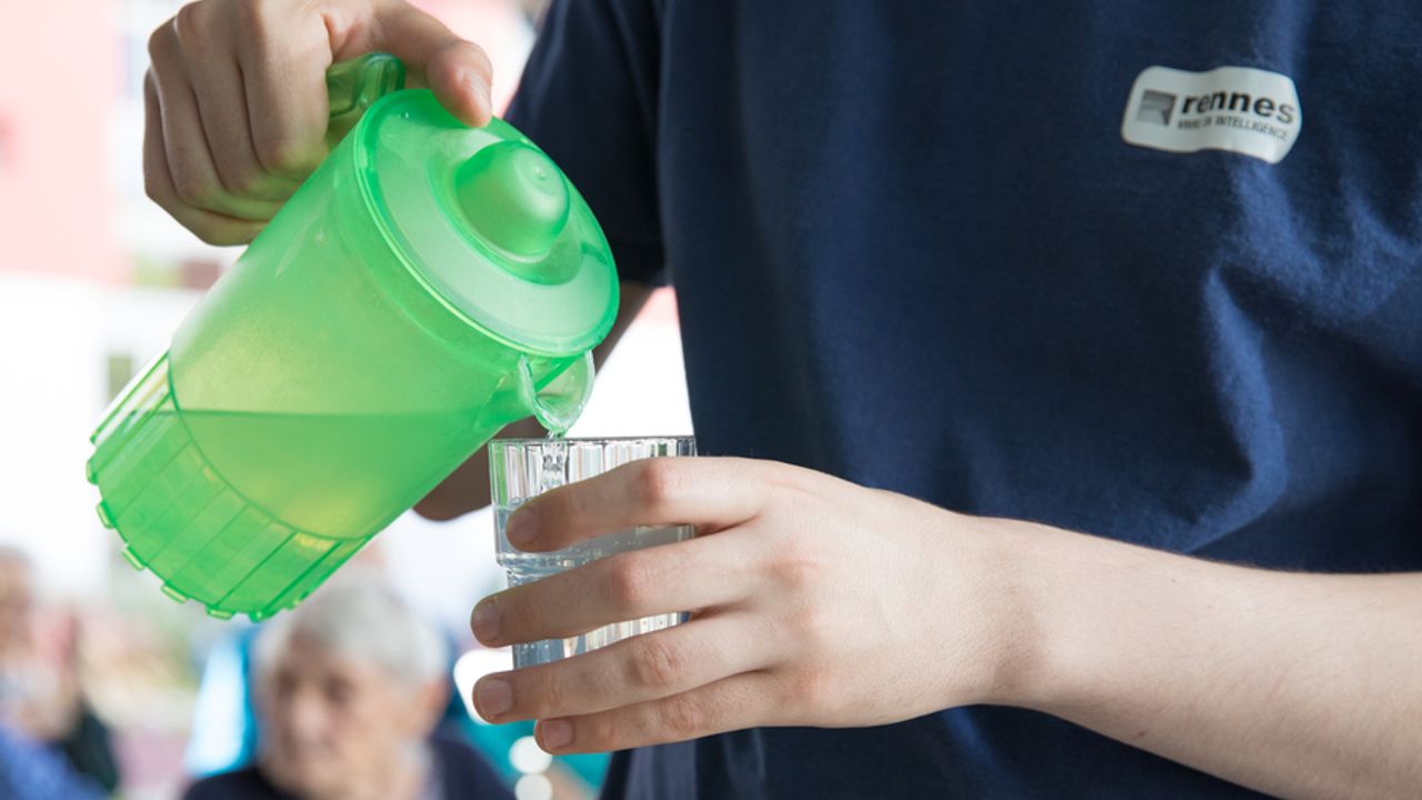 Une personne verse de l’eau dans un verre avec une carafe verte.