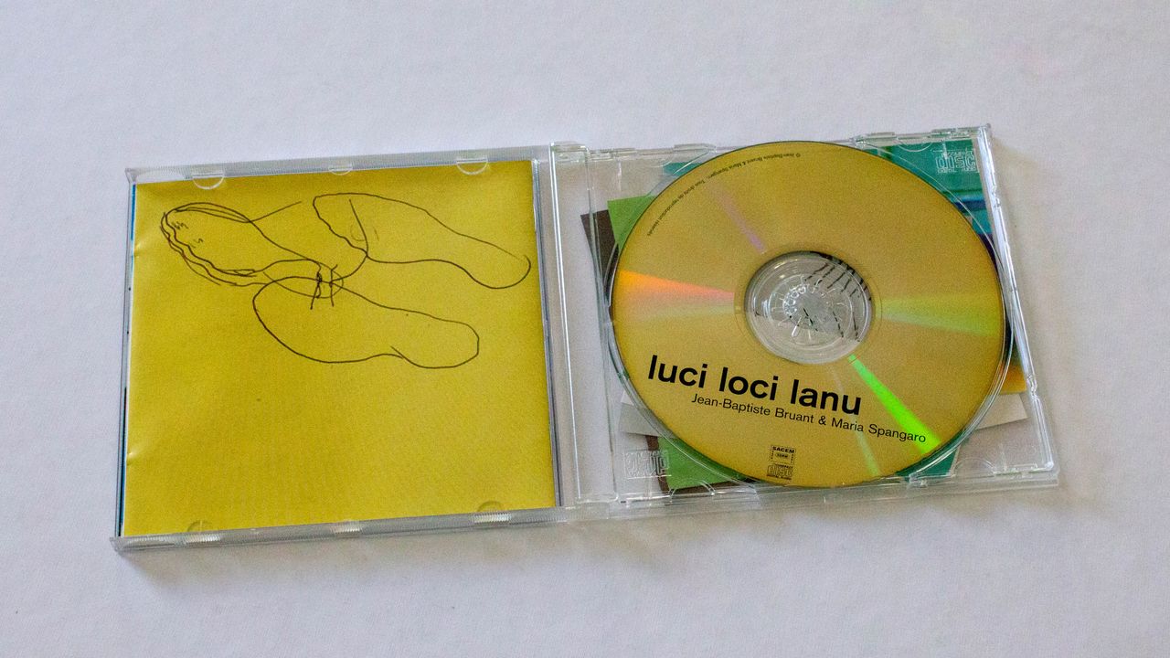 Boitier du cd ouvert. A gauche l'arrière du livret, jaune sur lequel on voit un dessin à main levé au stylo noir (on ne lit pas clairement un forme). A droite, le cd doré sur lequel on peut lire "luci loci lanu" puis en plus petit "Jean-Baptiste Bruant et Marie Spangaro"
