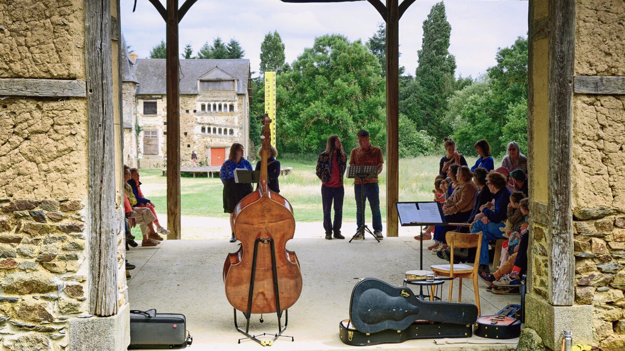 On aperçoit par une fenêtre du manoir de Tizé à Thorigné-Fouillard (dans l'agglomération de Rennes), une animation musicale. Au premier plan on voit l'arrière d'un violoncelle, un étui de guitare ouvert, une chaise, des feuilles posées sur un pupitre. Au seconde plan il y a des personnes (adultes et enfants) assises sur des chaises à gauche et à droite. Au troisième plan il y a 4 personnes debouts devant des pupitres qui discutent. A l'arrière plan on voit la fin du manoir et le mètre jaune XXL ainsi que des arbres et une étendue d'herbes.