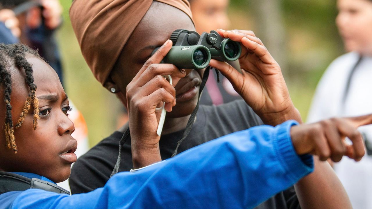 Deux enfants dans la nature : l'une pointe son index pour indiquer un oiseau, l'autre l'observe à travers des jumelles.