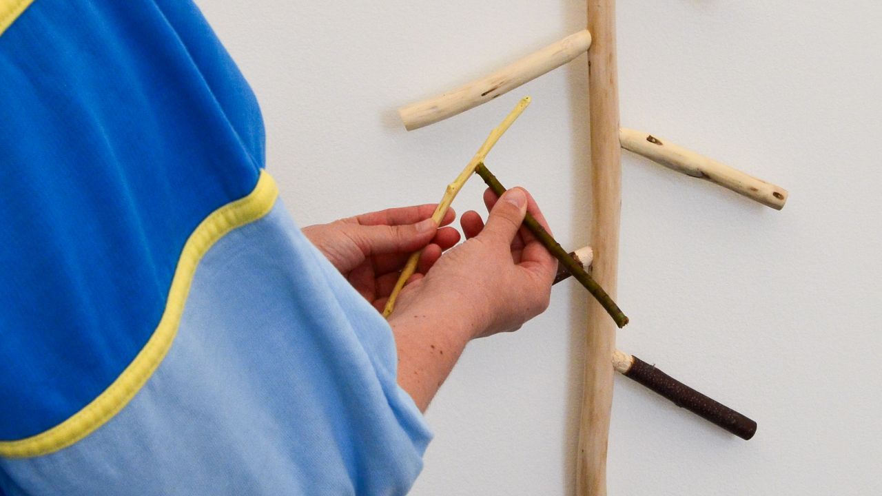 Une personne vêtue d'une veste bleue tient deux petits bâtons près d'une sculpture en bois fixée au mur et comportant plusieurs branches assemblées.