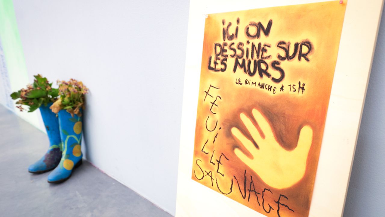 Un panneau indique "ici on dessine sur les murs, le dimanche à 15h" et "feuille sauvage". À côté, des bottes en caoutchouc d'où débordent des fleurs.