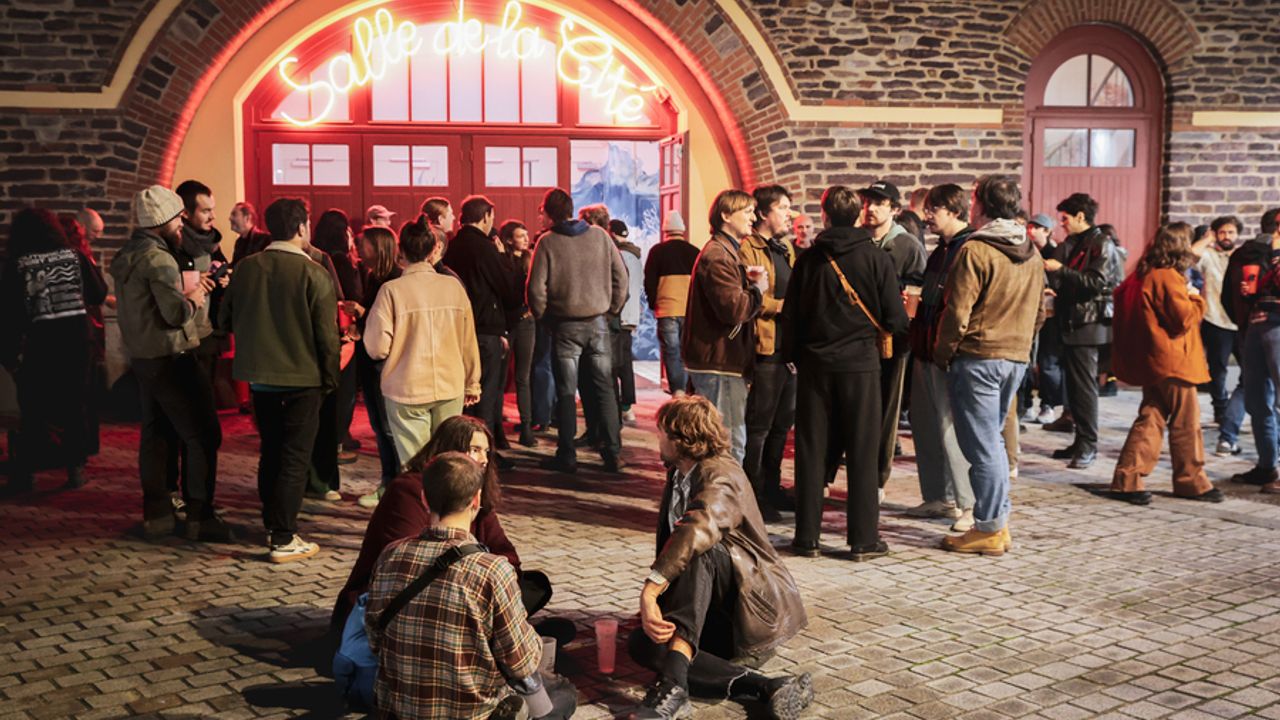 Vue de l’extérieur de la Salle de la Cité. Des personnes sont assises pendant que d’autres discutent debout devant l’entrée sous le néon de la salle.