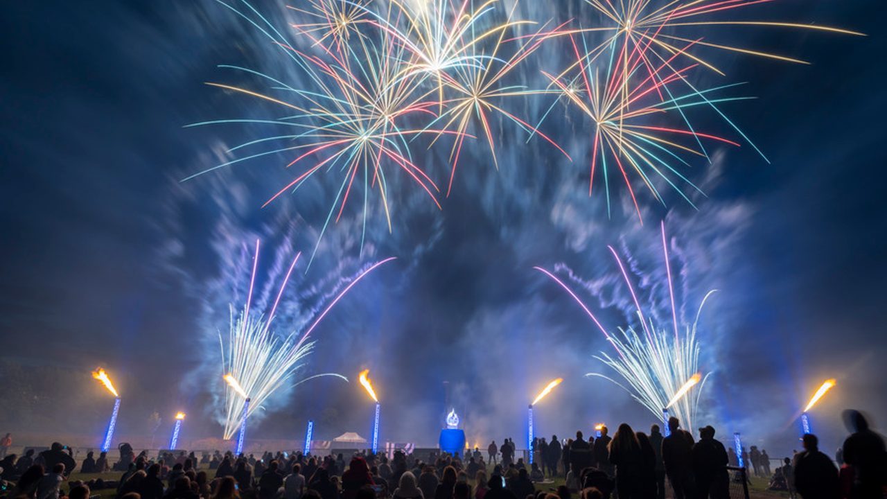 De nombreux habitants regardent le feu d'artifice au stade de la Bellangerais de Rennes le 13 juillet, une majorité des personnes est assise sur l'herbe. Dans le ciel on voit plusieurs artifices de différentes couleurs.