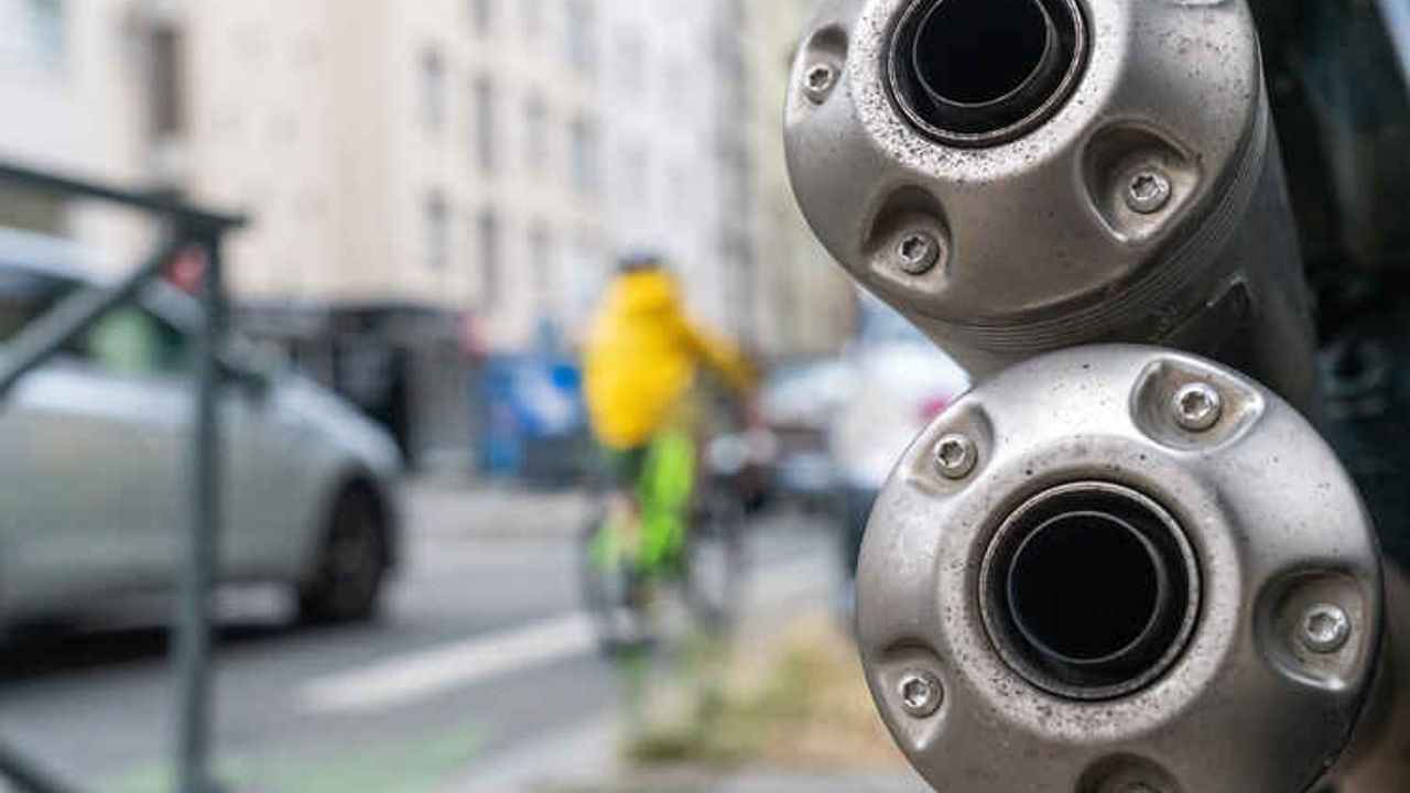 Pots d’échappement d’une moto en gros plan, dans une rue où circulent des voitures et un vélo.
