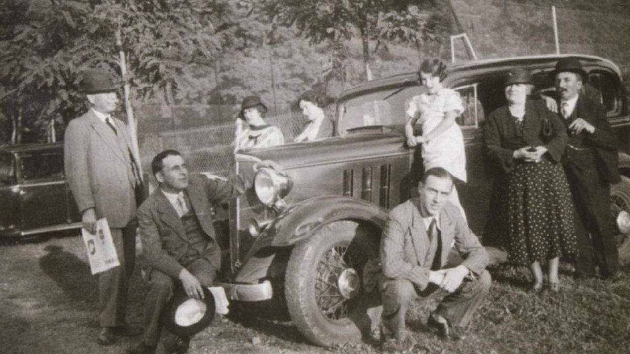 Plusieurs hommes et femmes posent devant une voiture. La scène se passe dvers 1930.