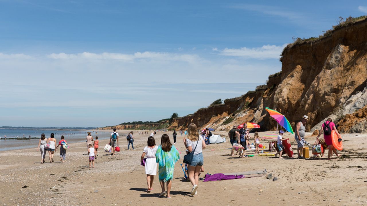 Sur la plage, au 1er plan 3 jeunes femmes s'avancent. En arrière plan, des personnes s'installent avec parasols