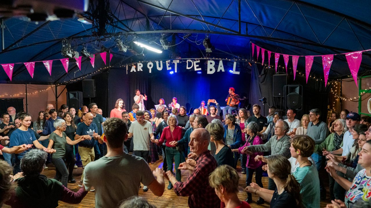 Des gens de tous les âges dansent dans une salle décorée par des fanions. À l'arrière plan, le bagad.