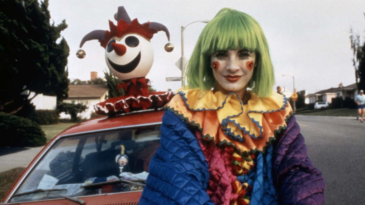 Au premier plan, une personne déguisée en clown avec des cheveux verts. Derrière elle, un joker avec des grelots sort du toit d'une voiture.