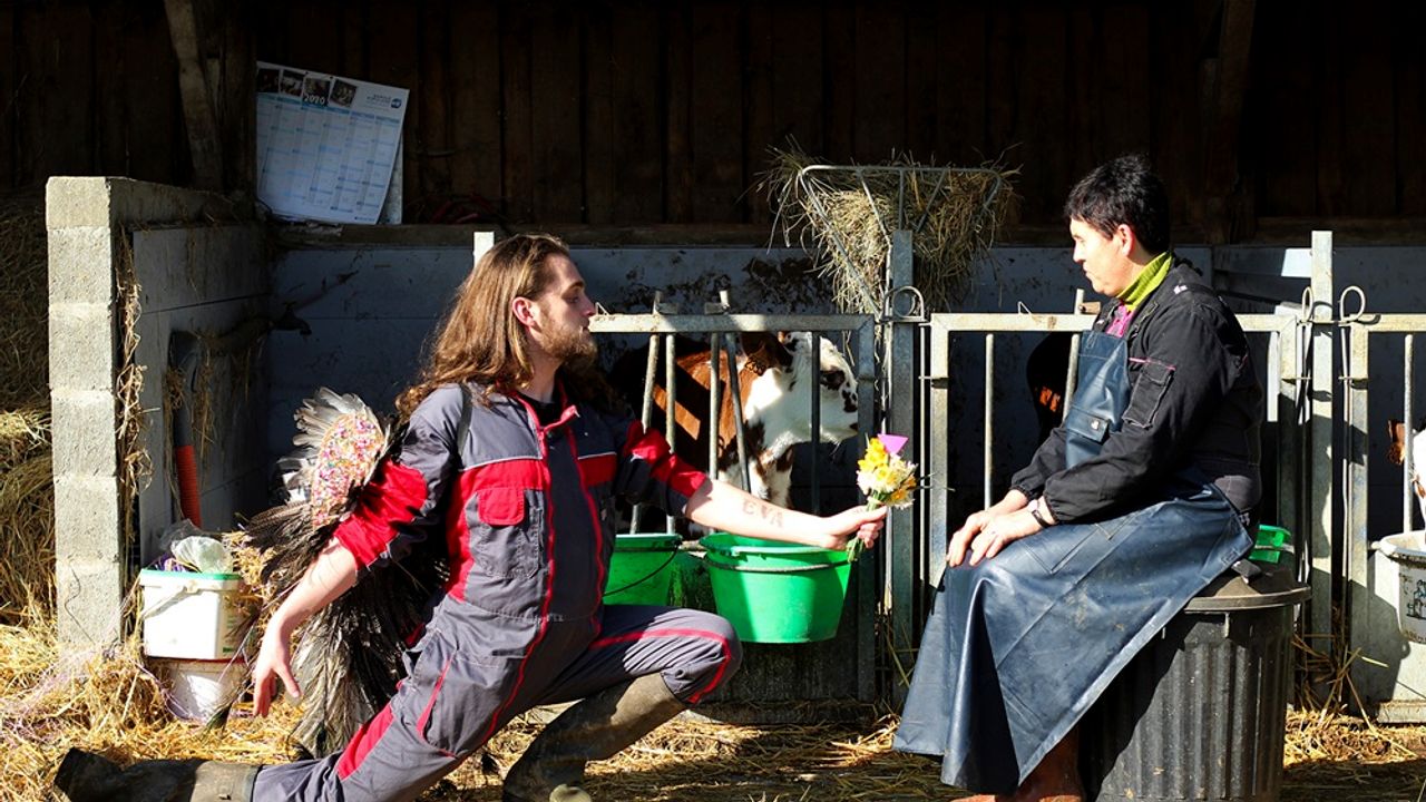 Dans une ferme, devant une stabulation, un hopmme au cheveux longs offre un bouquet de fleurs à une femmes assise sur une poubelle. Tous les deux sont en tenue ed travail, il y a de la paille au sol.