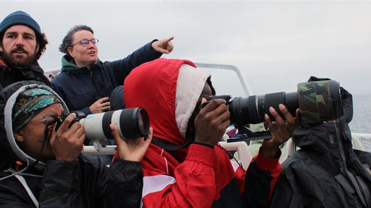 À bord d'un bateau, des jeunes ajustent leurs objectis pour prendre des photos, accompagnés par des animateurs.