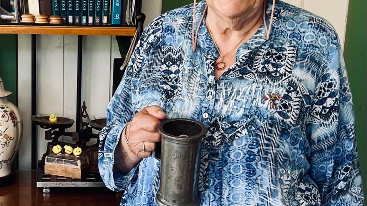 Monique vêtue d'une chemise bleue pose devant un meuble avec bibelots. Elle a dans sa main, un pot d'étain qui servait à mesurer le vinaigre dans son épicerie