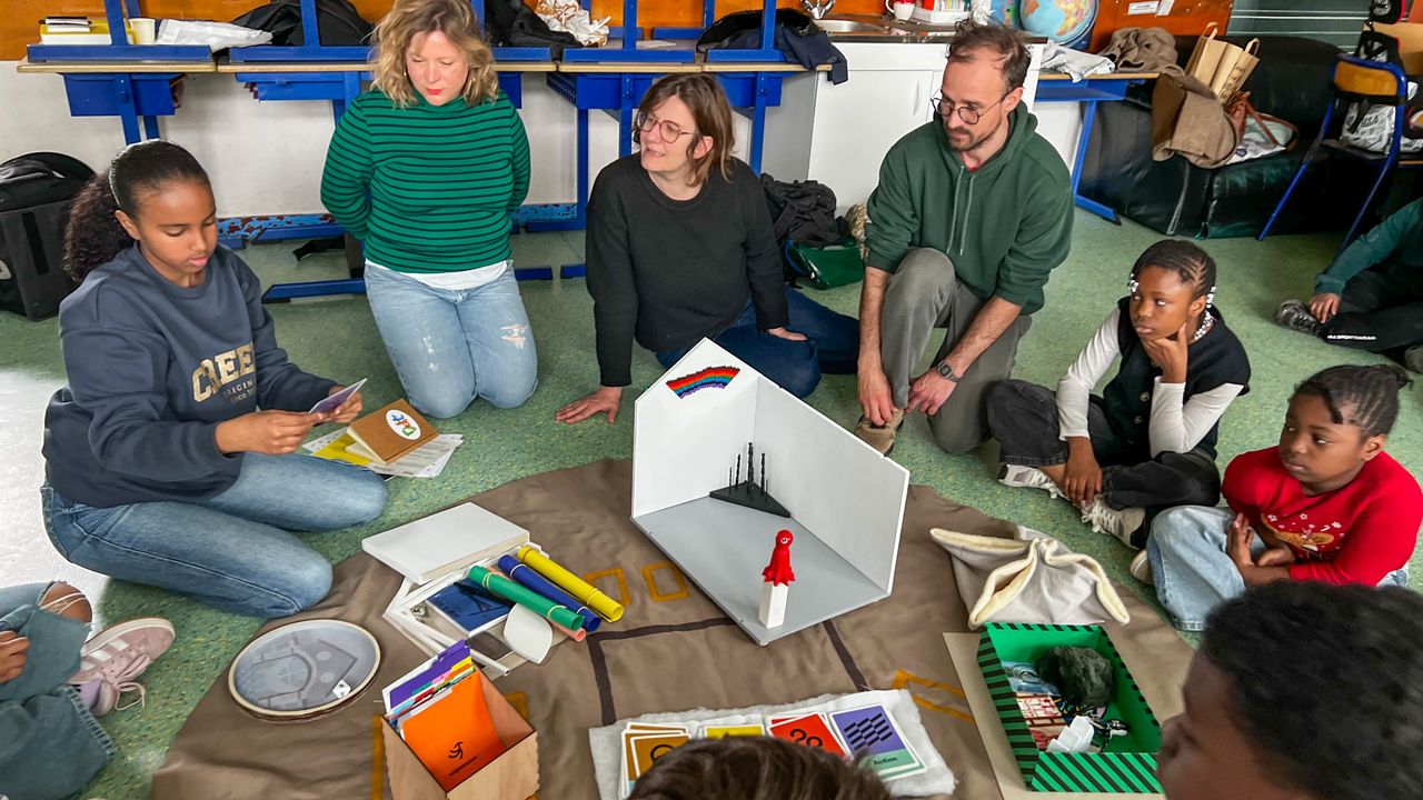 Des enfants ainsi que les artistes Fanny Martel et Yann Baïzid, et la médiatrice Amandine Braud, sont assis par terre en rond autour du tapis de jeu du prototype du jeu Rejouer La Criée. Une enfant lit une carte au reste de la classe.