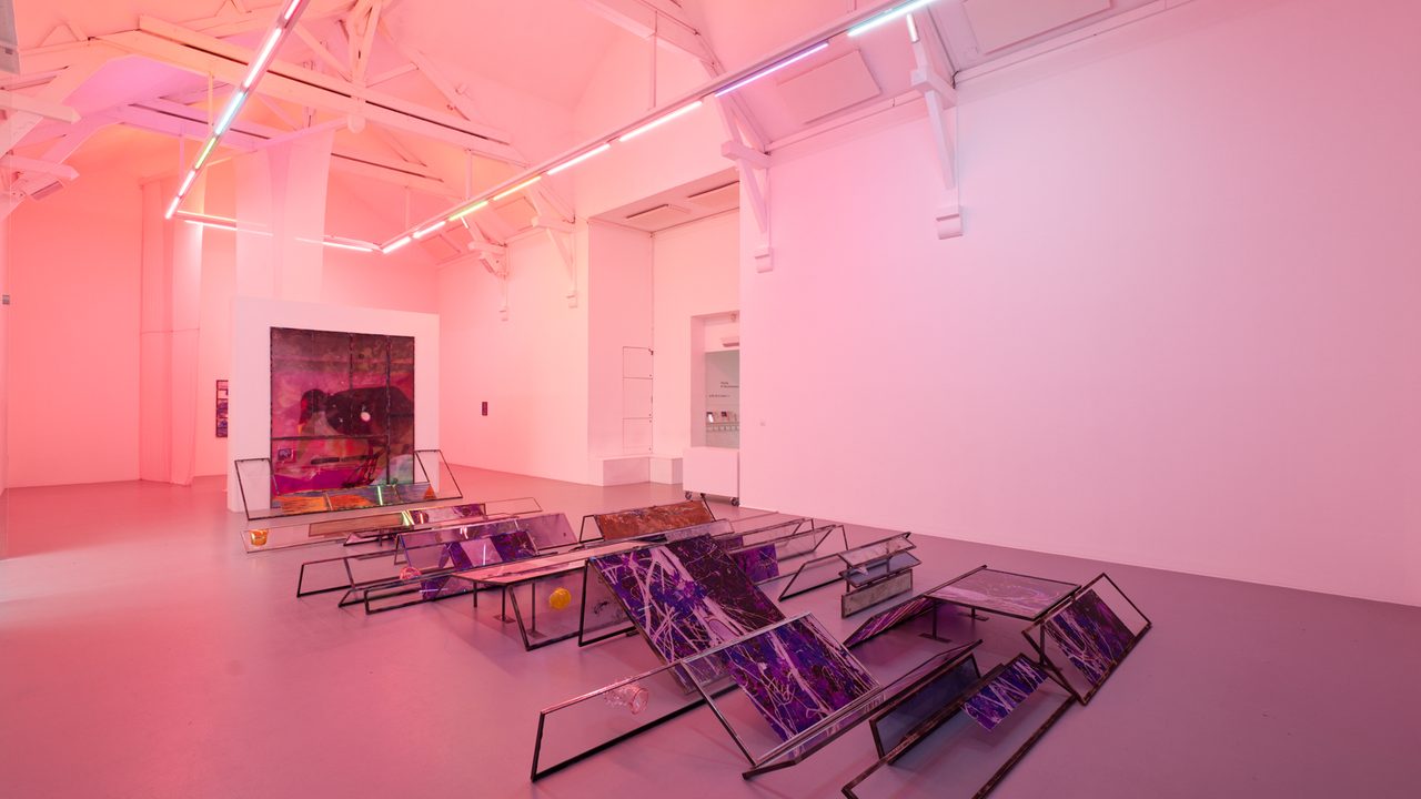 Salle d’exposition avec au sol l’œuvre d’Euridice Zaituna Kala. Des panneaux de verre au sol et des néons au plafond.