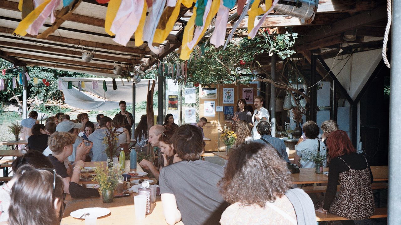 Crowd of people sitting around tables set outside on a porch. There's colourful decorations hanging and people are eating