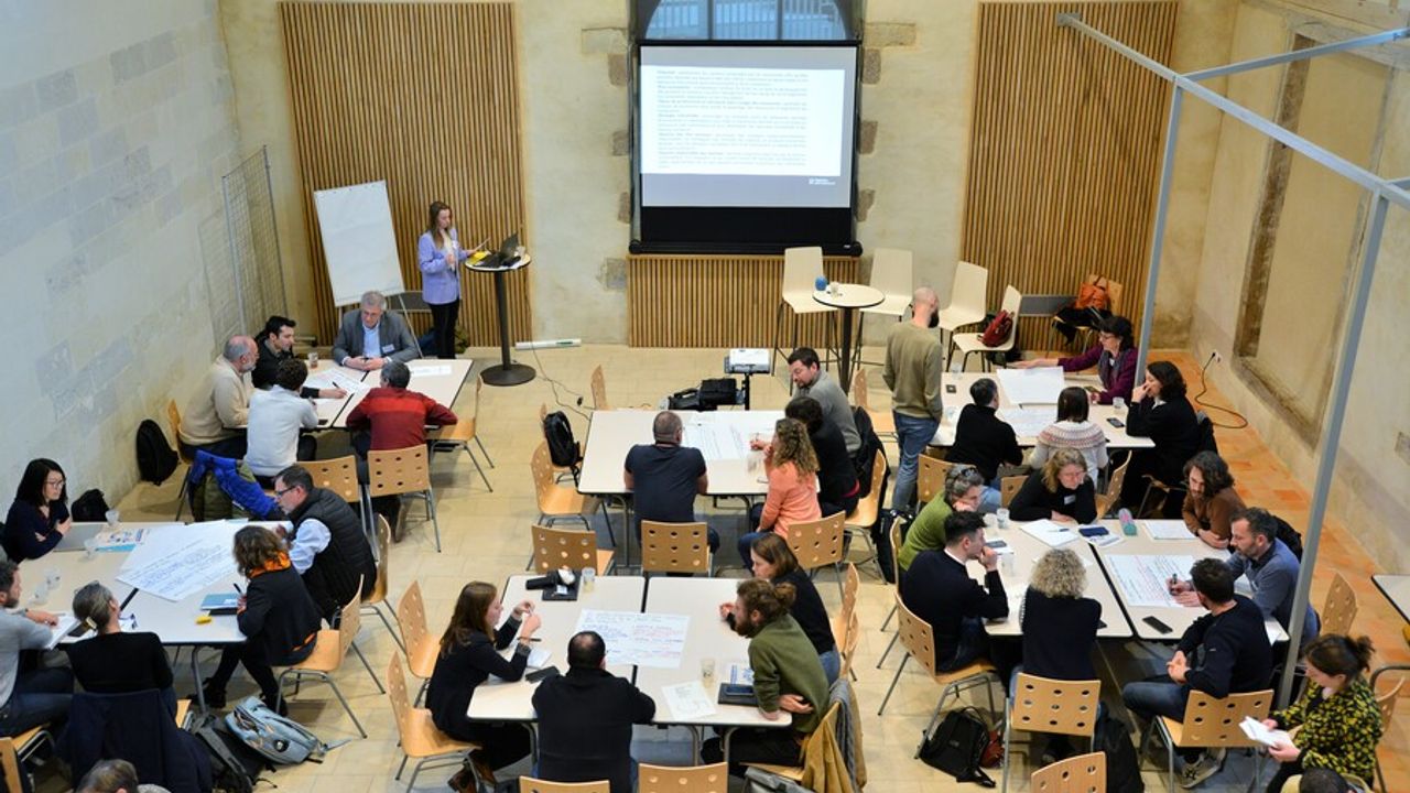 Des personnes attablées travaillent en petits groupes. Vue de l’intérieur de la salle du Jeu de Paume.