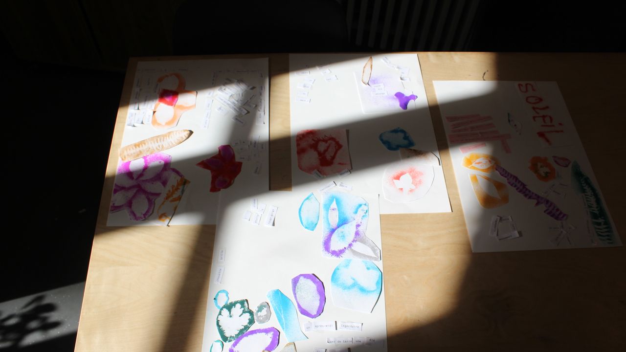 A la lumière du soleil les feuilles A3 colorées composées par les élèves sont réparties sur une table en bois