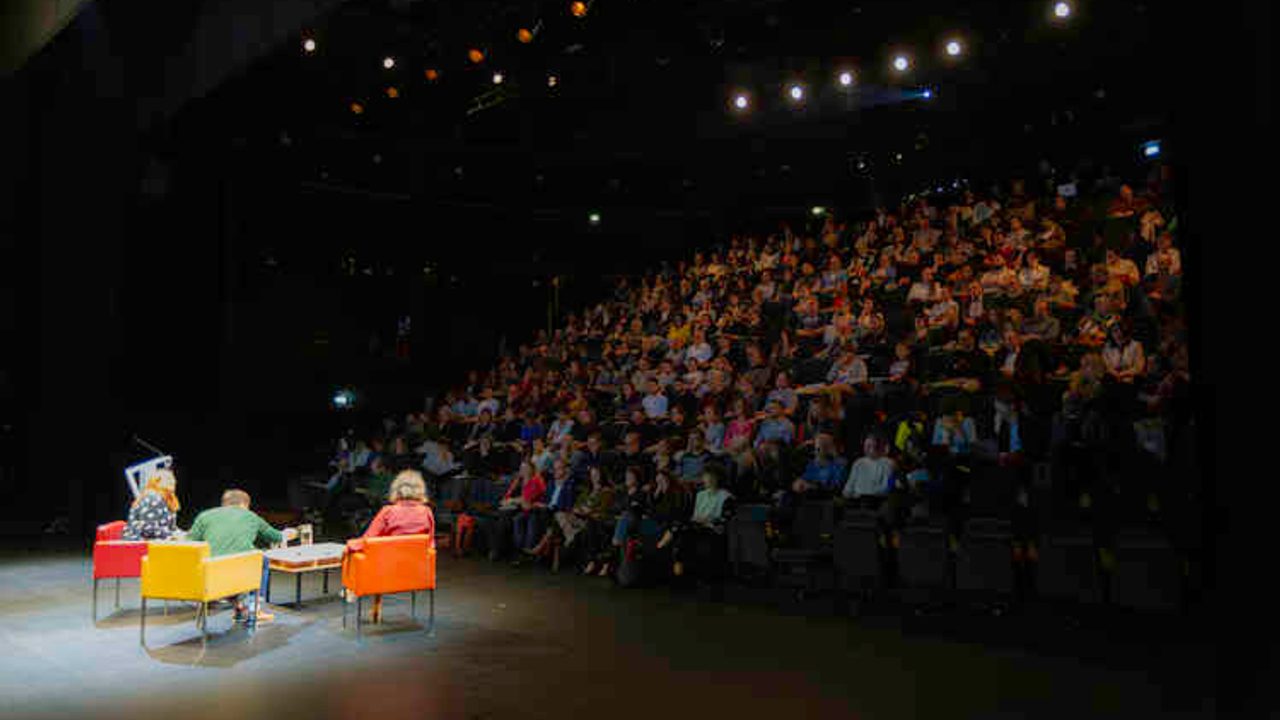 salle de conférence remplie avec trois intervenants sur scène