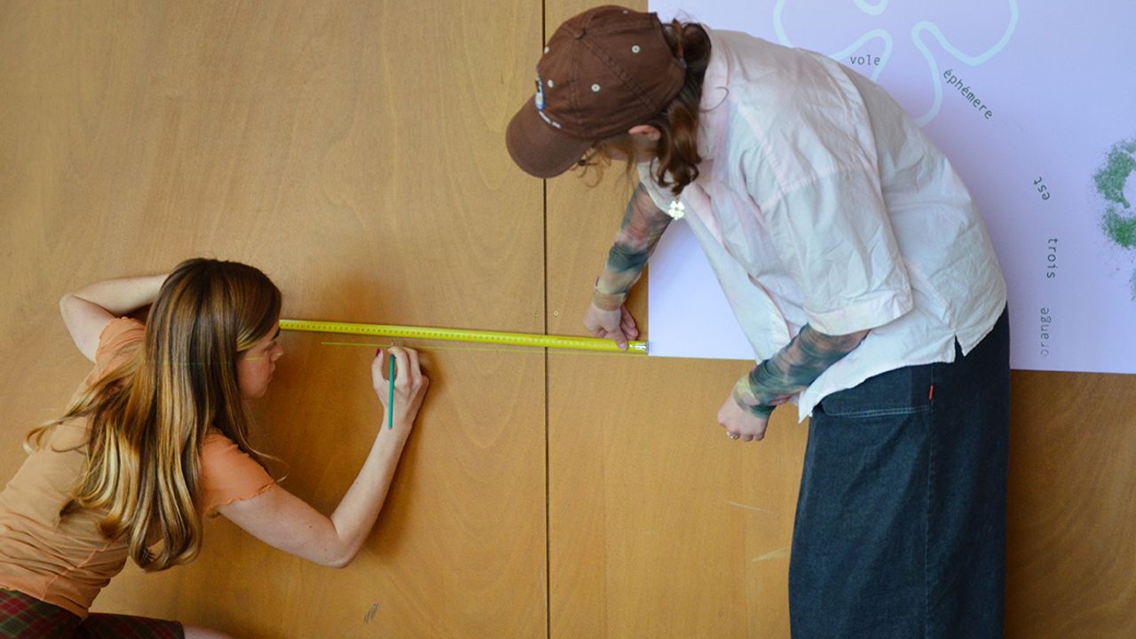 Les artistes Lucie Férézou et Margaux Janisset accrochent leurs affiches sous le préau de l'école.