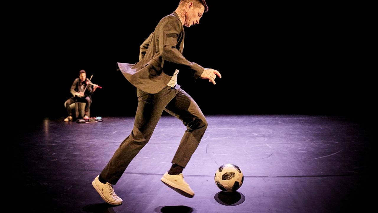 Au premier plan, un homme en costume joue au football sur une scène, avec un violoniste au second plan