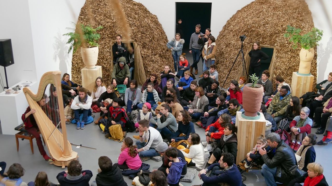 Public écoutant un concert de harpe au milieu d'oeuvres d'art en forme d'œufs, nids d'oiseaux.