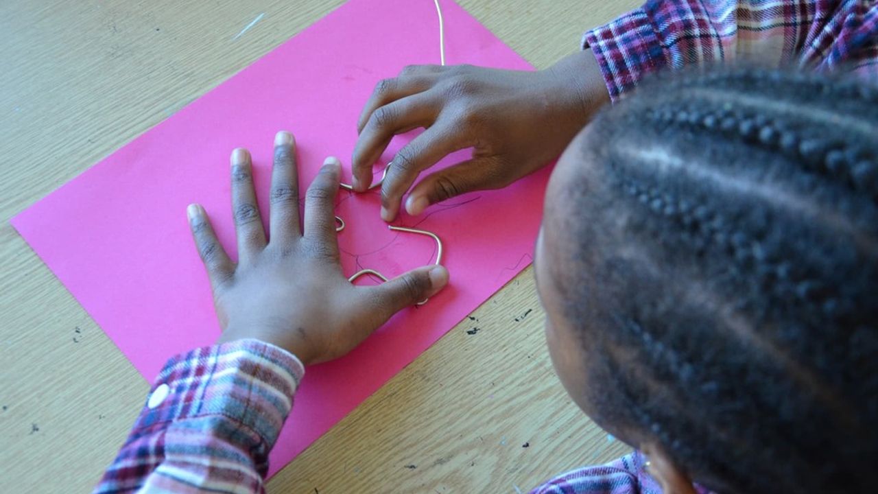 On peut observer un enfant vu du dessus en train de fabriquer une feuille de chêne avec un fil sur un papier rose.