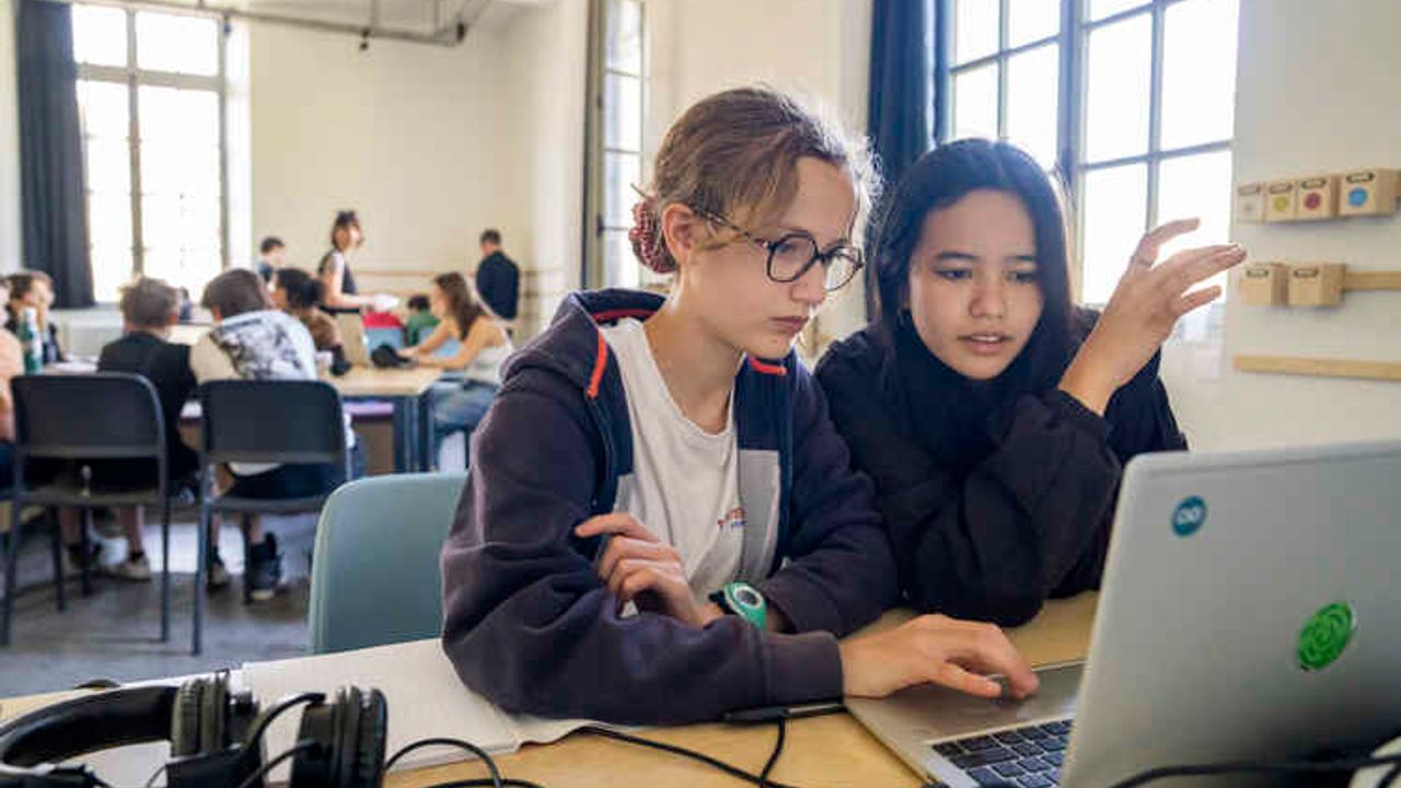 deux jeunes filles sont concentrées devant un ordinateur, dans une salle de classe