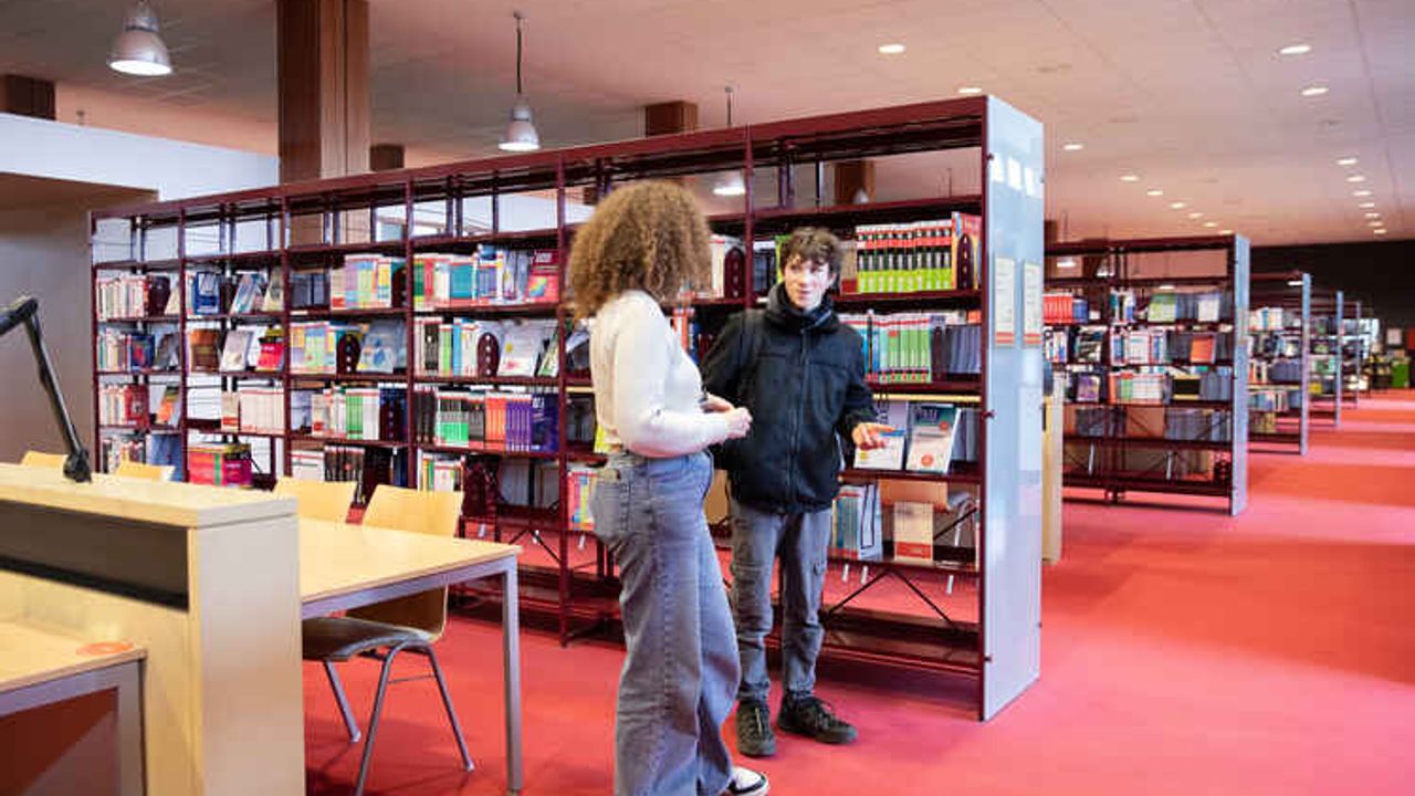 Dans la salle de lecture d’une bibliothèque universitaire, une femme et un homme discutent debout au centre de la photo. La femme est tournée de côté vers l’homme qui est de face. Derrière lui, des rayonnages de livres en enfilade jusqu’à l’arrière-plan. Sur la gauche de la photo, une table et quatre chaises vides.
