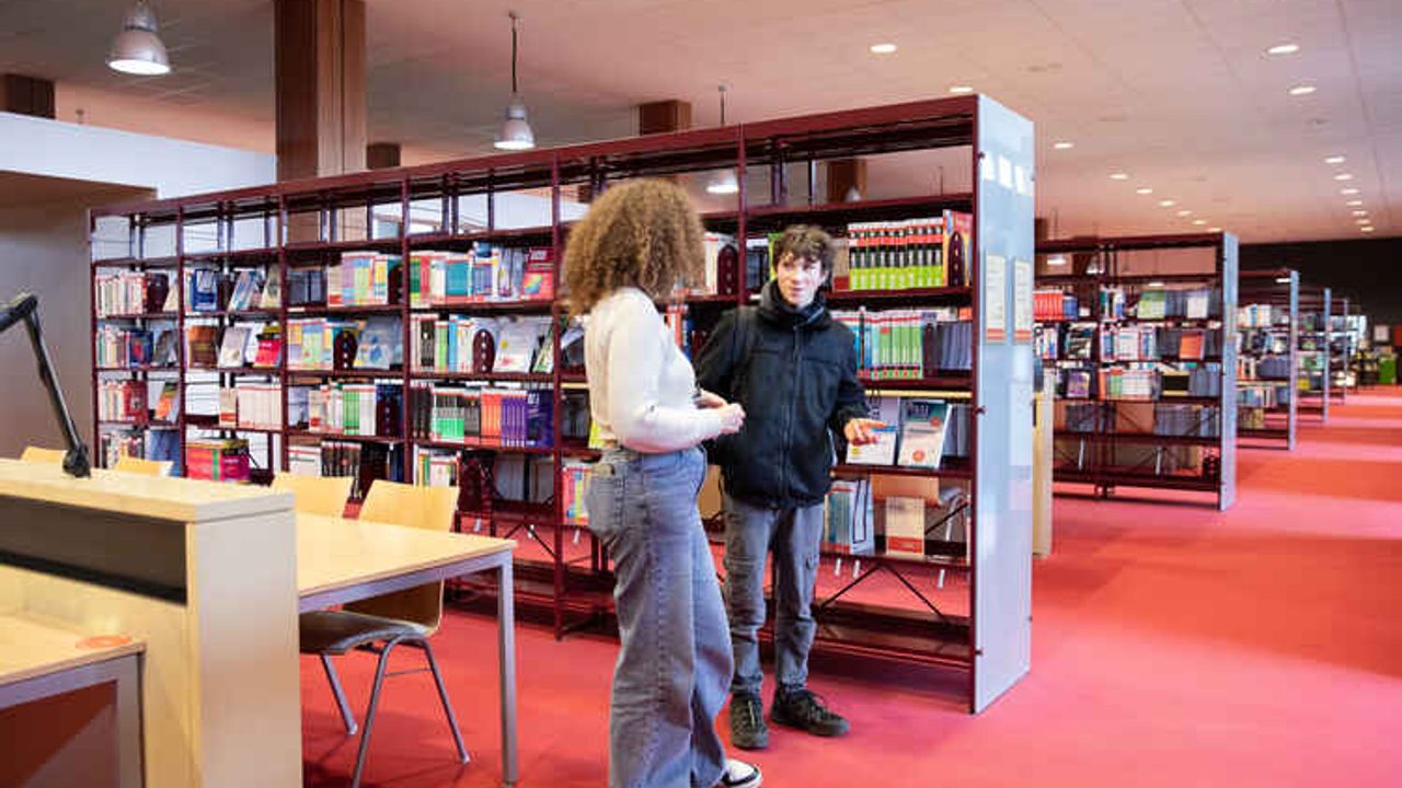 Dans la salle de lecture d’une bibliothèque universitaire, une femme et un homme discutent debout au centre de la photo. La femme est tournée de côté vers l’homme qui est de face. Derrière lui, des rayonnages de livres en enfilade jusqu’à l’arrière-plan. Sur la gauche de la photo, une table et quatre chaises vides.