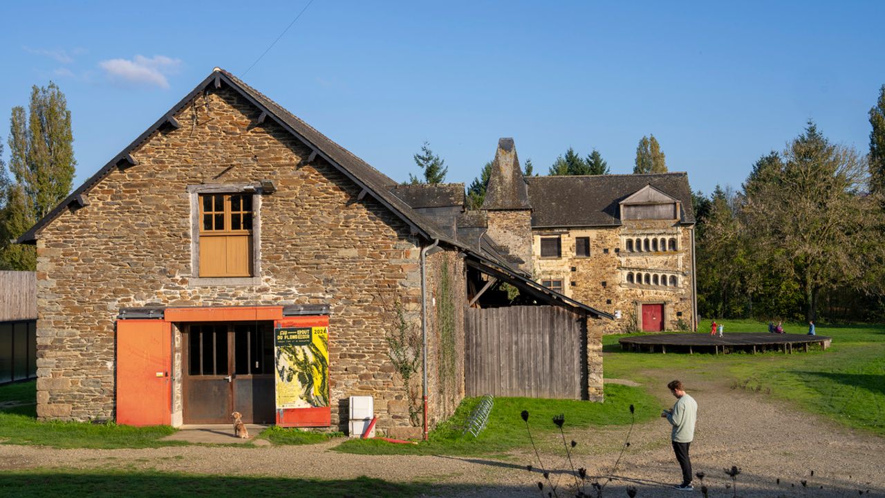 C'est le manoir du domaine de Tizé à Thorigné-Fouillard dans l'agglomération de Rennes (35), l'association Au bout du plongeoir occupe les lieux et organise des événements accessibles au public. On voit le bâtiment en pierre avec une affiche de l'association à droite de la porte d'entrée. Il y a une personne et un chien devant l'entrée. Au fond on aperçoit plusieurs personnes dans le jardin. Derrière le bâtiment, il y a de nombreux arbres. Le ciel est bleu.