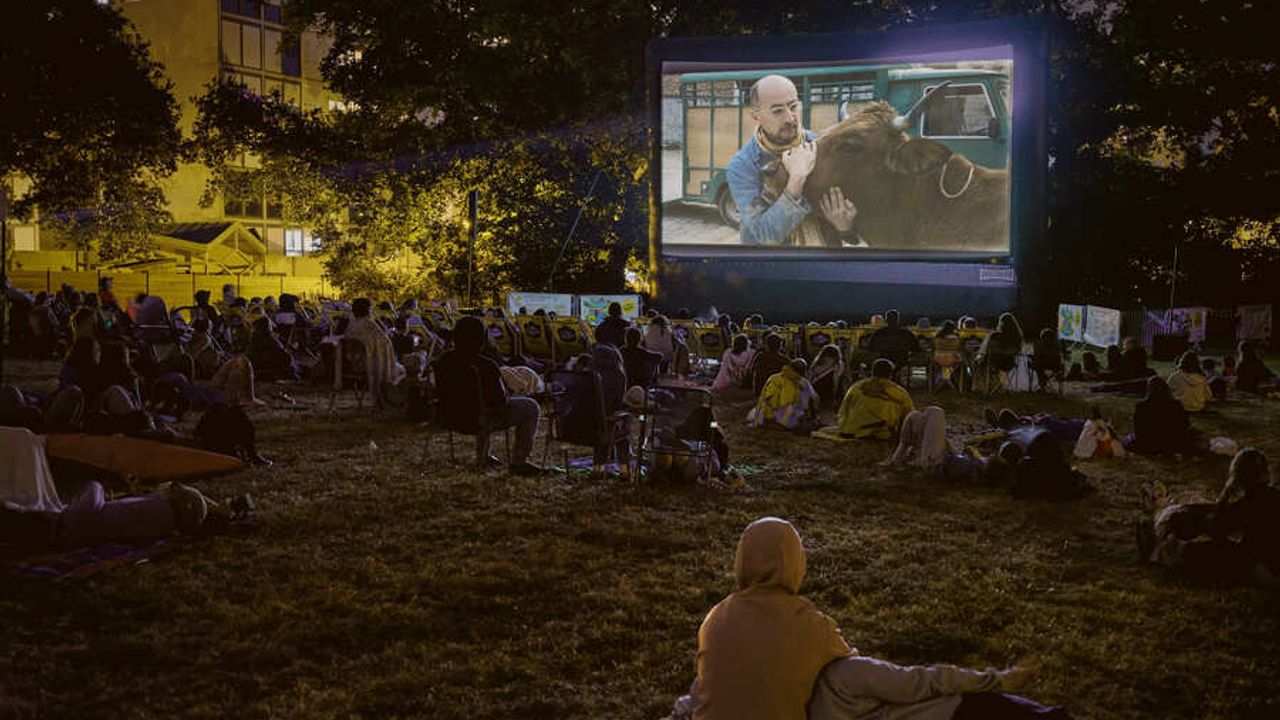 A la nuit tombée, des personnes sont assises par terre de dos à regarder un écran diffusant un film où l'on voit un agriculteur et sa vache.