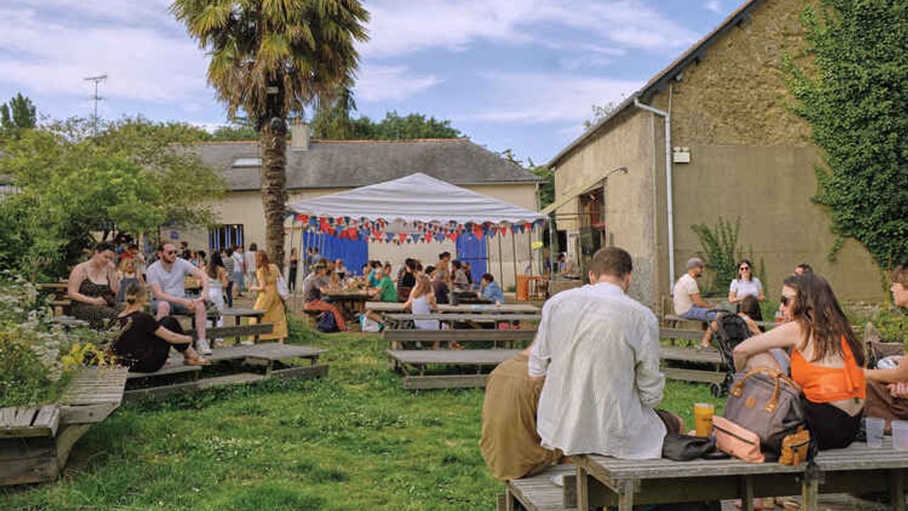 Sur la pelouse, une ginguette avec des tables et fanions. Les personnes par groupe discutent.