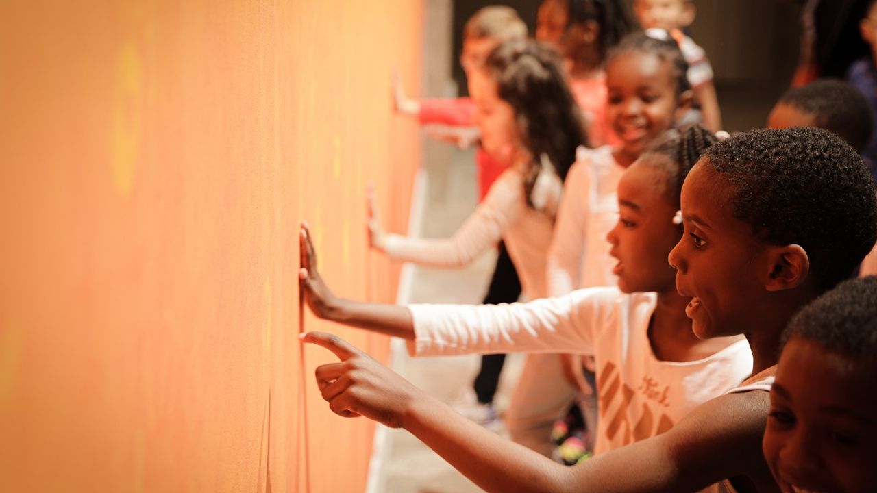 Des enfants découvrent une exposition en touchant un mur de couleur orangé.