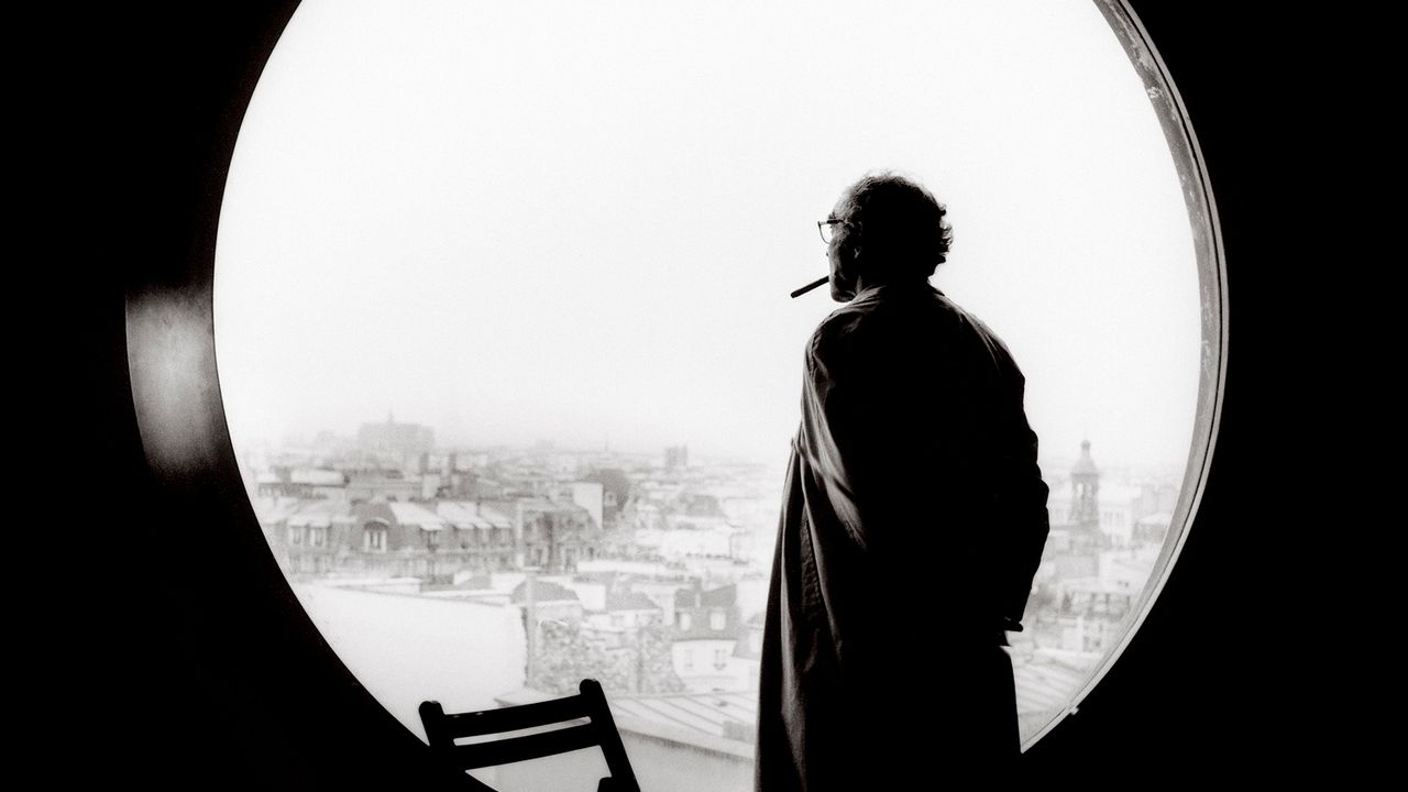 Le réalisateur Jean-Luc Godard de dos, regarde la ville à travers une fenêtre ronde