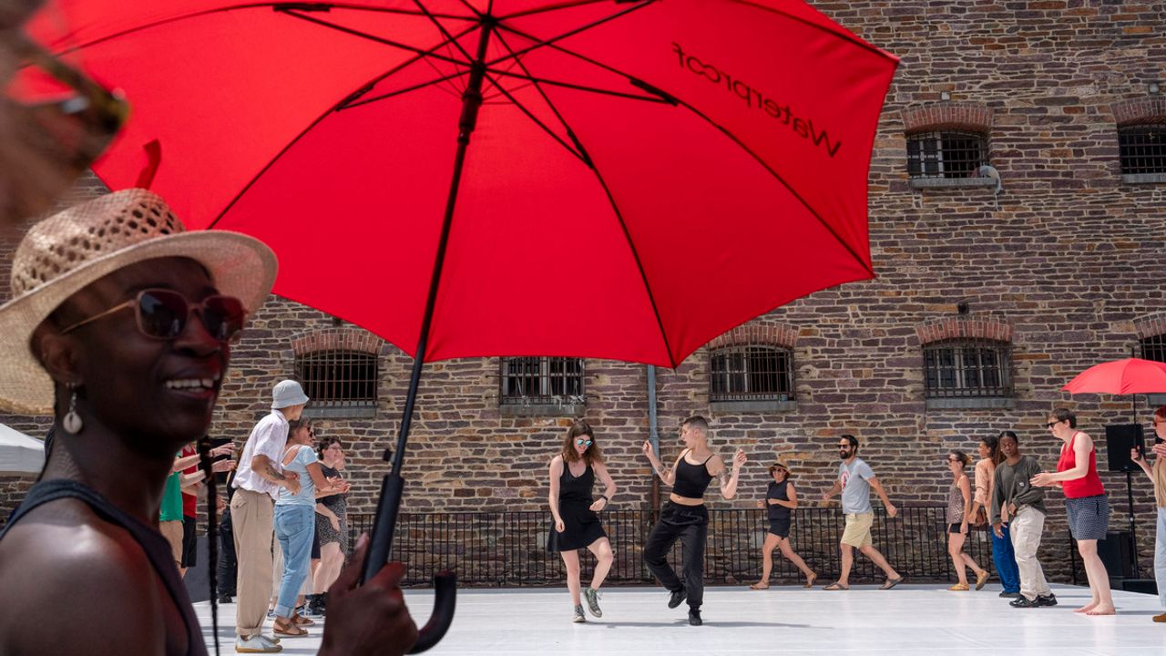 Au premier plan, une femme qui tient un parapluie sous le soleil, et en arrière fond, des danseurs contemporains.