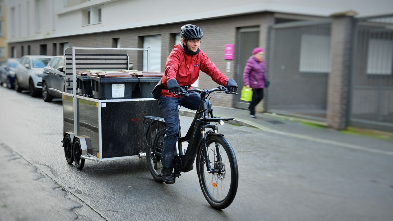 Une cycliste pilote un vélo avec une remorque dans laquelle sont rangées des poubelles.
