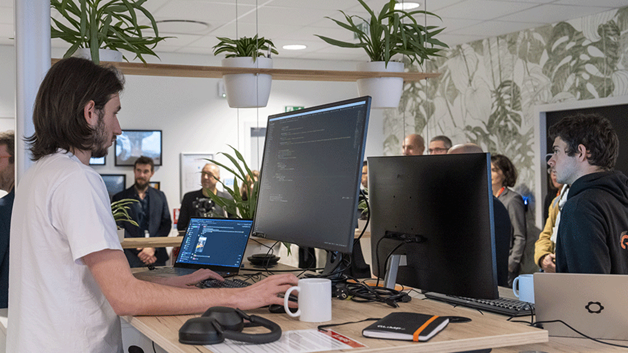 Un ingénieur informatique au premier plan avec ses écrans d'ordinateur. Au fond, des personnes visitent la cyberplace.