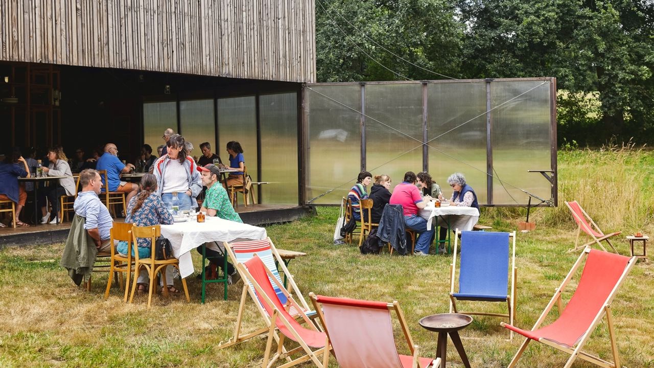 On voit la terrasse du manoir de Tizé à Thorigné-Fouillard (35) occupé par l'association Au bout du plongeoir. Terrasse ouverte sur l'extérieur avec une partie abritée comprenant des tables et chaises ; une partie sur l'herbe comprenant des tables et chaises et transats. Plusieurs groupes de personnes sont assis.