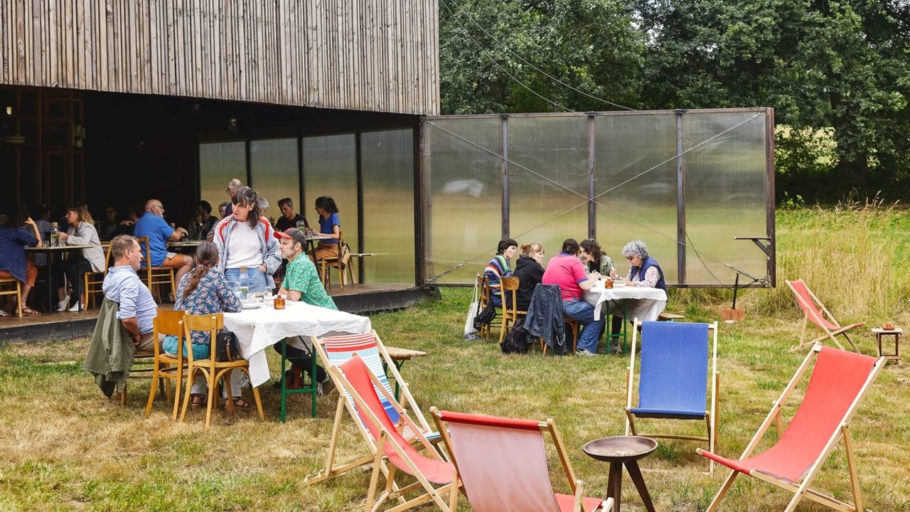 On voit la terrasse du manoir de Tizé à Thorigné-Fouillard (35) occupé par l'association Au bout du plongeoir. Terrasse ouverte sur l'extérieur avec une partie abritée comprenant des tables et chaises ; une partie sur l'herbe comprenant des tables et chaises et transats. Plusieurs groupes de personnes sont assis.