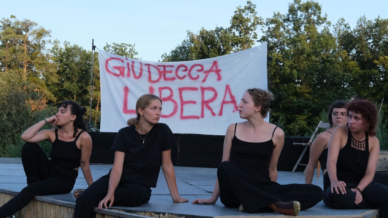 People performing at an outdoor stage at golden hour, with a banner saying "Giudecca libera"